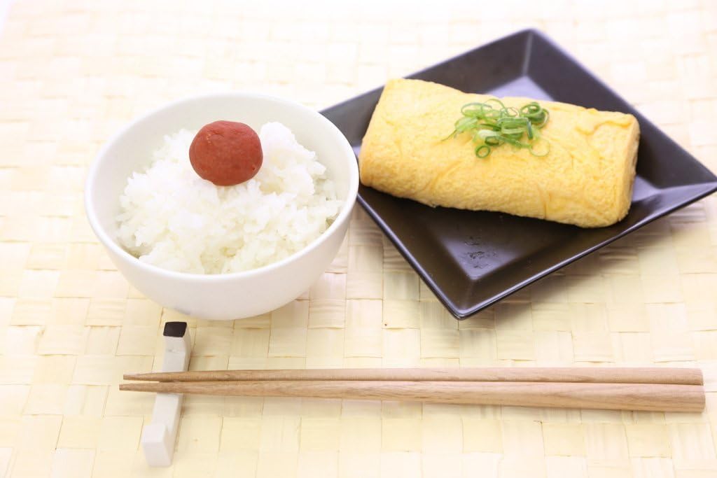 A finished rolled tamagoyaki topped with sliced spring onion on a black plate beside a bowl of rice
