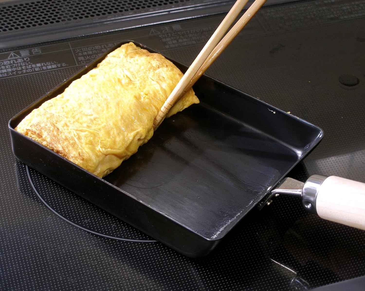 A rolled tamagoyaki being folded with cooking chopsticks in a River Light iron egg pan on an induction hob
