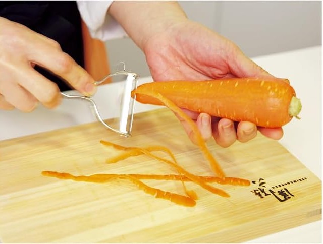 Kai Select 100 T-peeler taking thin strips of skin off a carrot over a wooden cutting board