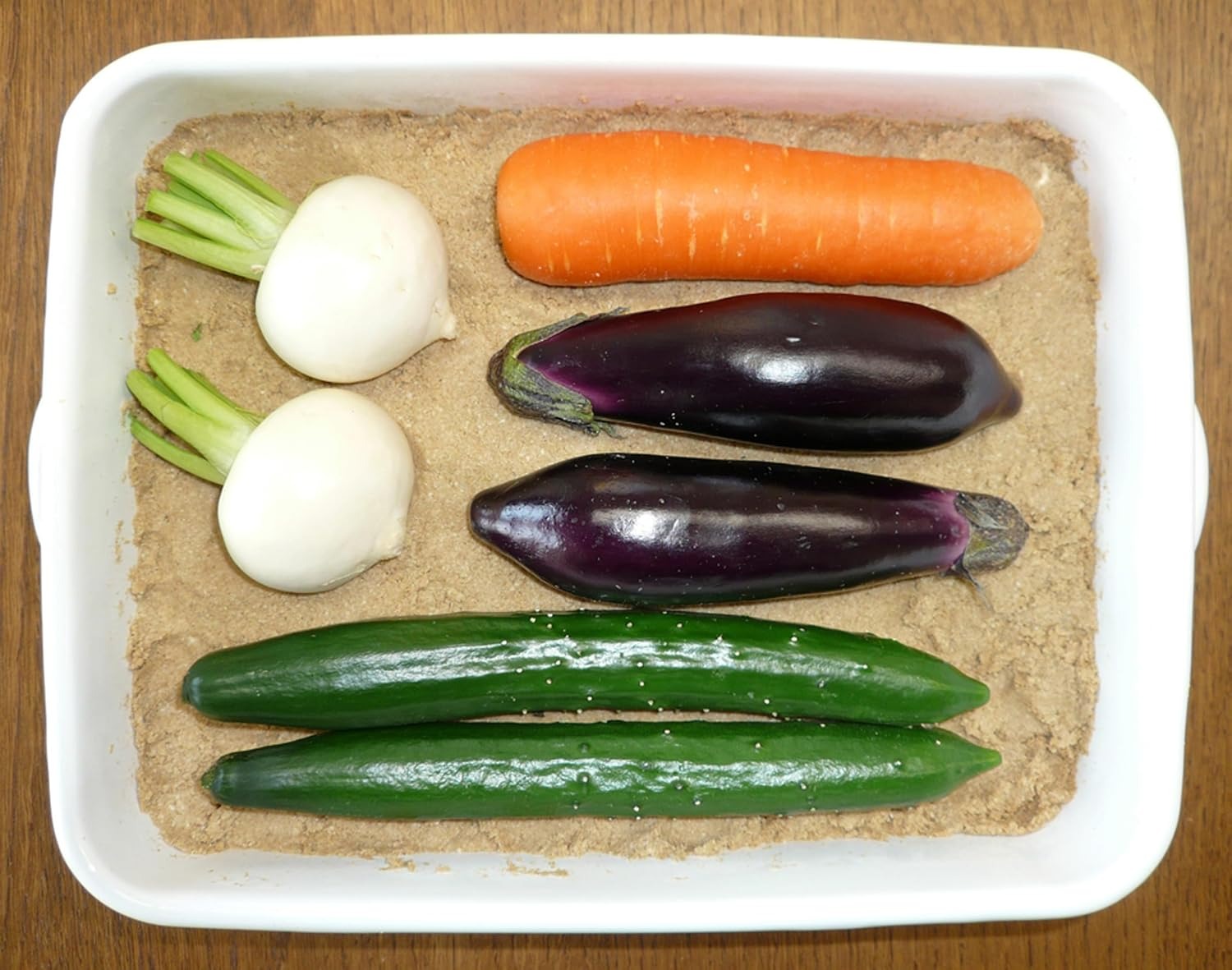 Cucumbers, eggplants, a carrot and small turnips resting on a rice-bran pickle bed inside a white enamel container