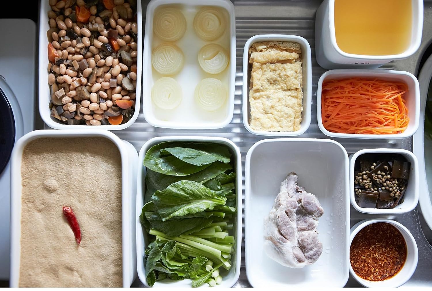 White enamel containers filled with prepped beans, greens, carrot, fried tofu and a rice-bran pickle bed